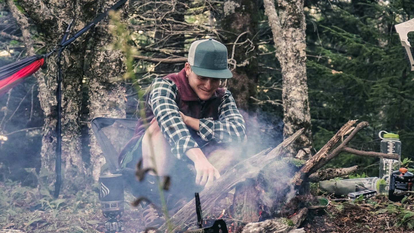 Man in a plaid shirt tends a campfire in the woods.