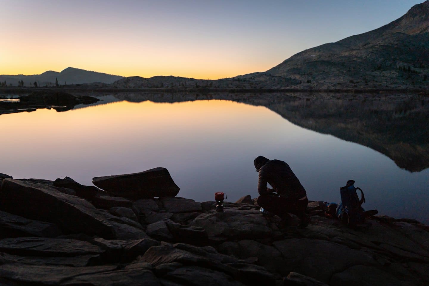 Person cooking on camping stove by mountain lake at twilight.