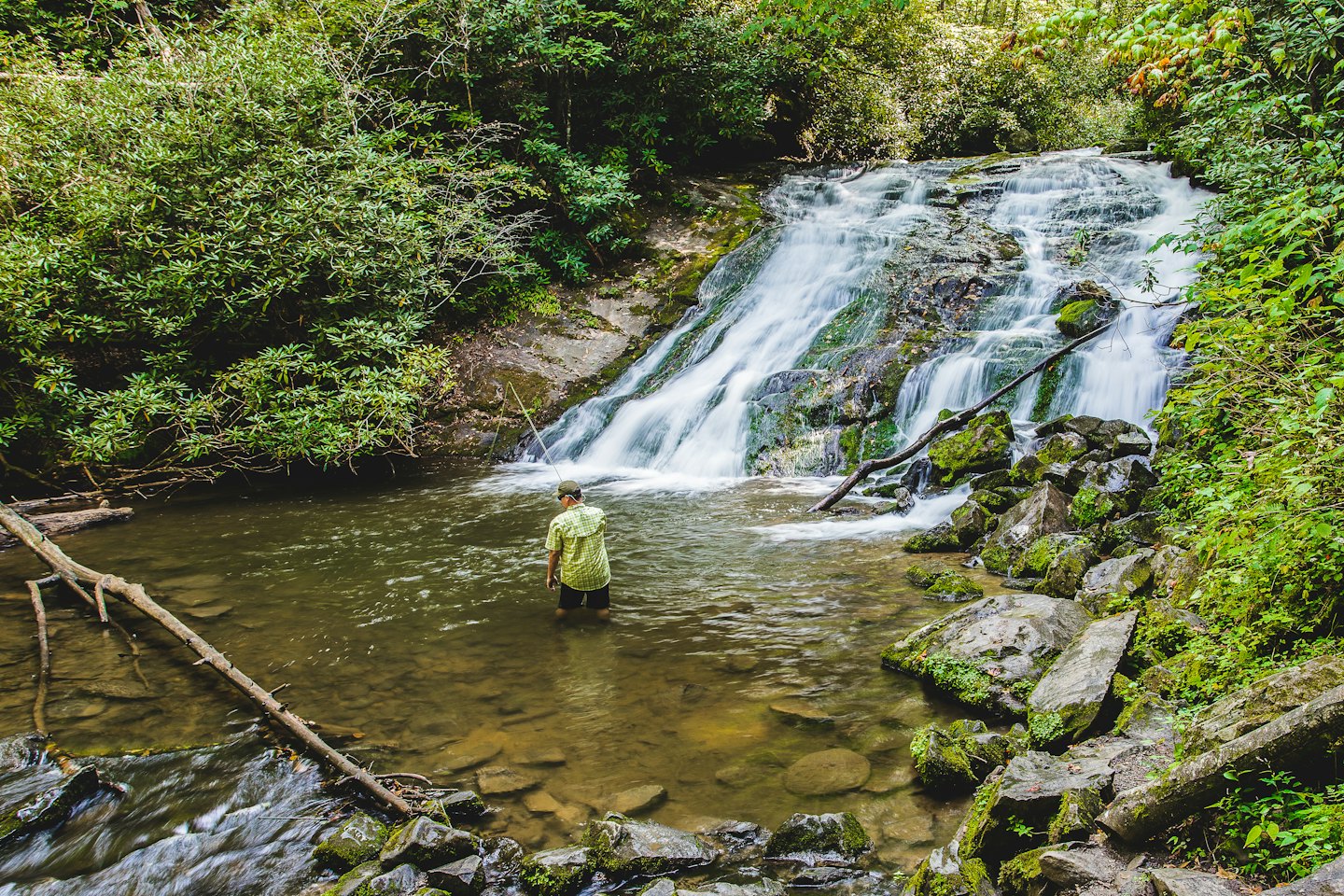 Fly fisher stands in a river before a tiered waterfall.