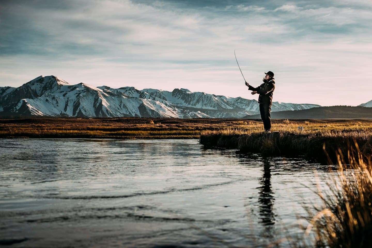 Man fly-fishing on a riverbank with snowy mountains.