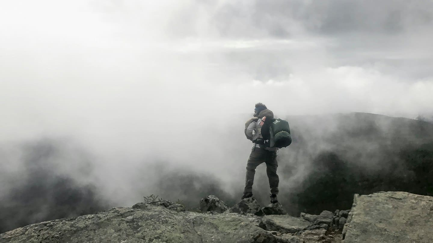 Backpacker standing on a rocky mountain peak overlooking misty valleys.
